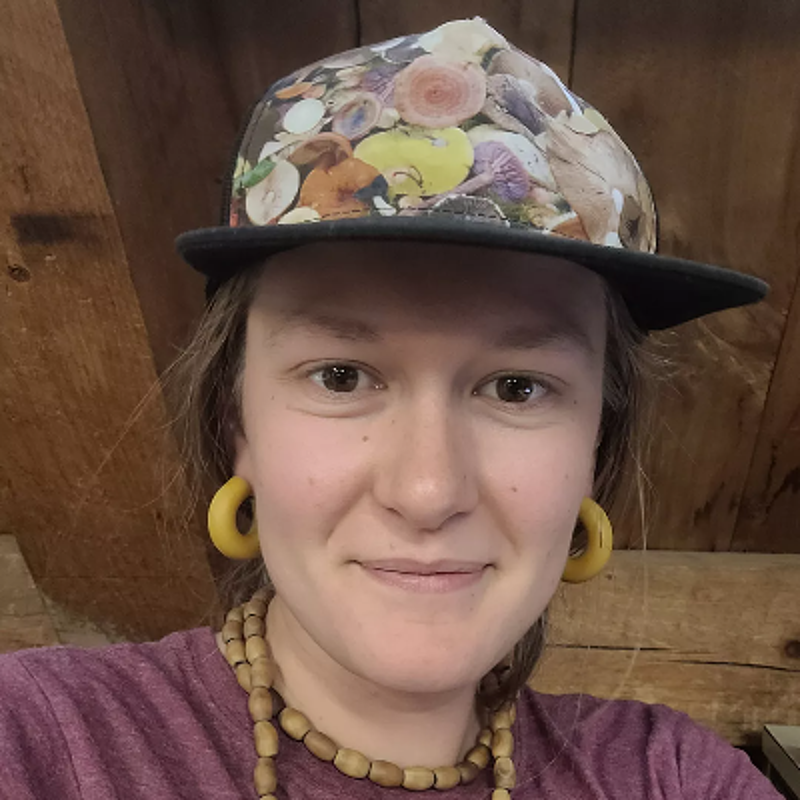 The image shows a person with fair skin, brown eyes, and long hair. They are wearing a hat with a colorful floral design, large yellow hoop earrings, and a maroon shirt. They are also wearing a beaded necklace. The background appears to be a wooden wall.